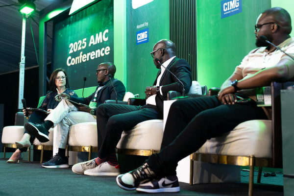Panelists seated on a stage during the 2025 AMC Conference panel discussion, with a woman speaking into a microphone on the left.