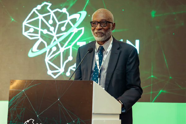 Bespectacled man in a navy suit speaks at a podium during a conference, with a neon network graphic backdrop.