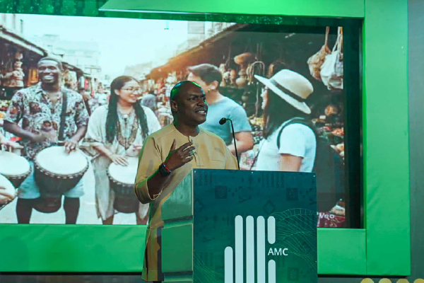 Man in a tan outfit speaks at a podium on a green-lit stage, with a large screen behind him showing drummers and shoppers.