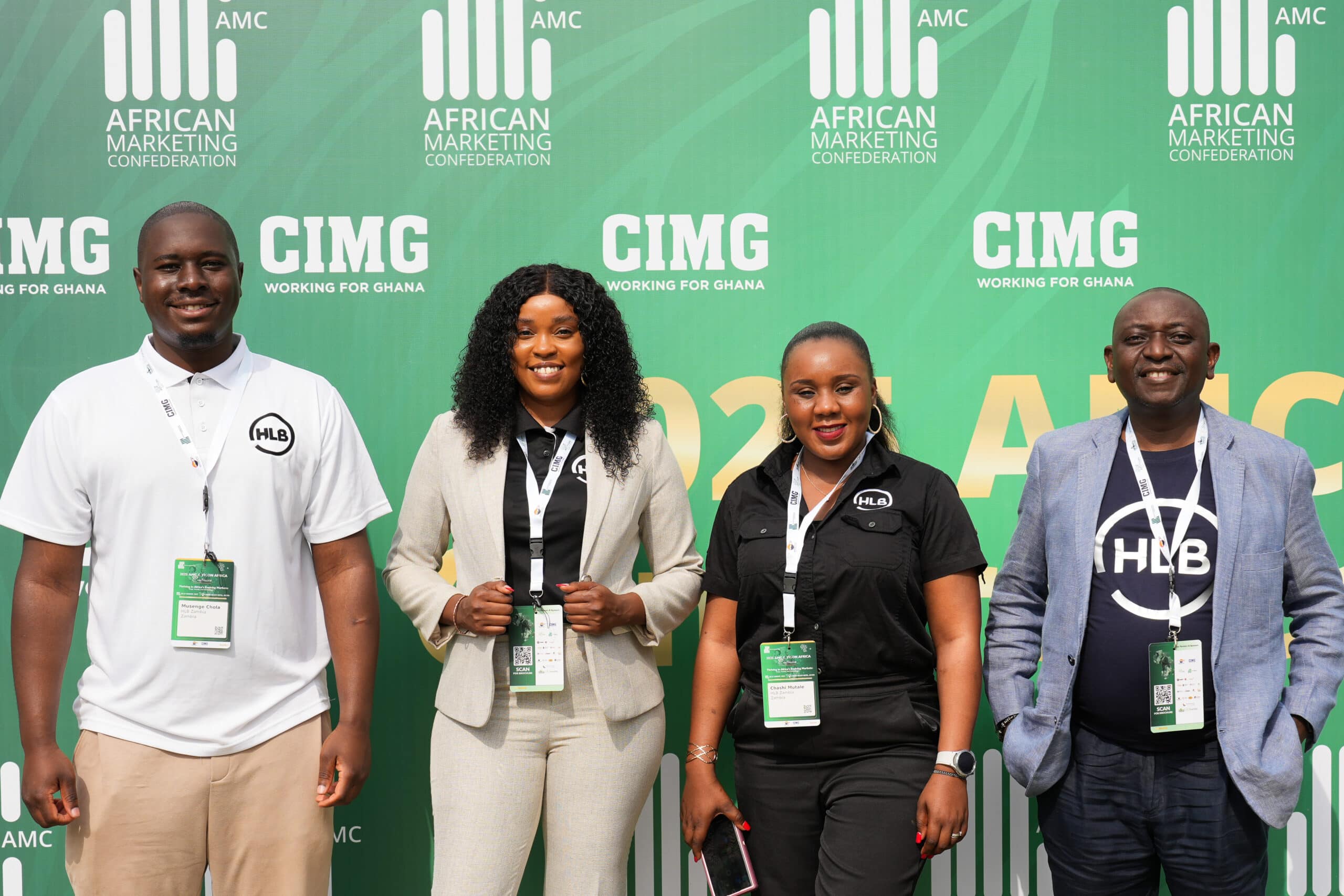 Four people stand in front of a green backdrop with AMC branding; from left to right: man in white polo with HLB badge, woman in light beige suit, woman in black shirt with lanyard, man in blue blazer and dark shirt.